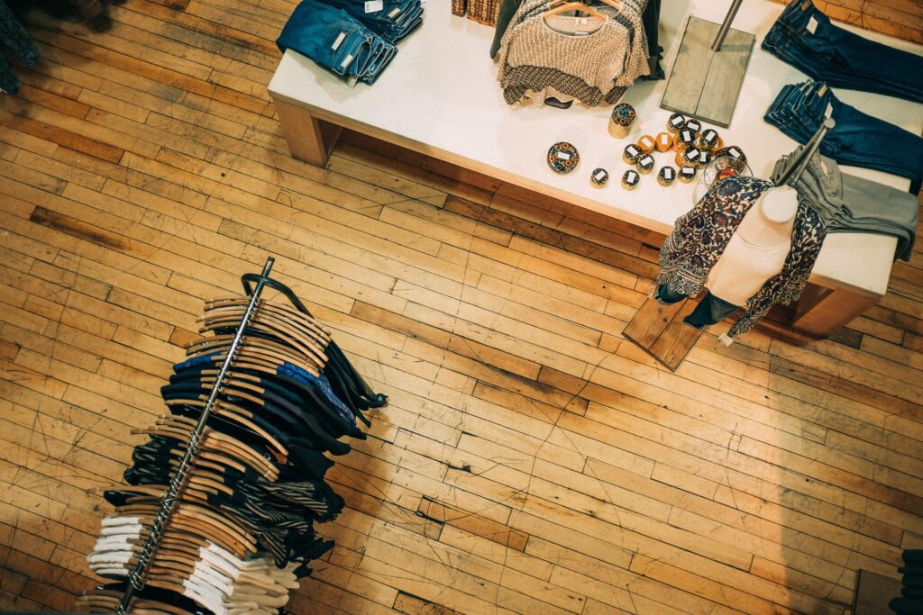 Clothing store interior showing a wooden floor, a round rack with hanging clothes, and a table displaying folded clothes and small bowls.