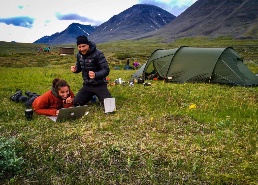 Two people in outdoor gear are on grass near a tent in a mountainous area. One lies on the ground using a laptop, while the other stands nearby with hands raised. Camping gear is scattered around.