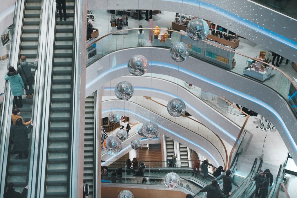 Modern shopping mall interior with multiple floors, escalators, and decorative hanging silver spheres. Shoppers are seen on the escalators and walking through the open, well-lit space.