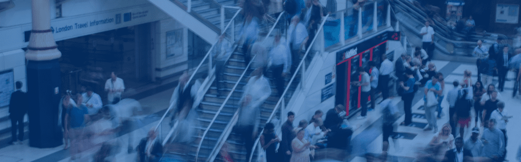 Blurred crowd moving up and down escalators and walking in a busy train station, with a few people standing near a kiosk.