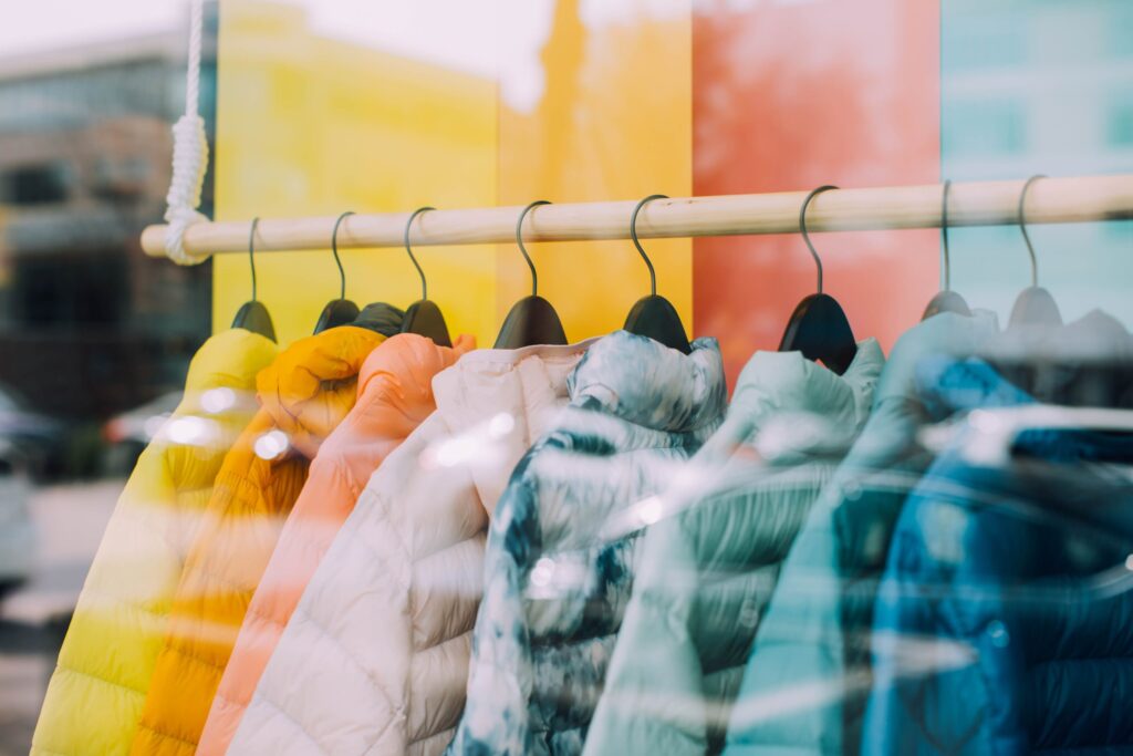 Several puffer jackets in different colors are hanging on a wooden rack in a store display, with a reflection of the street visible in the window.