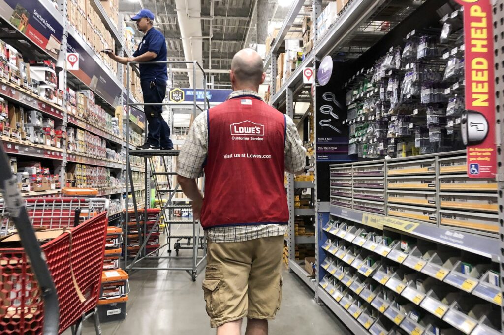 A Lowe's employee in a red vest walks down an aisle as another worker stocks shelves in a hardware store.