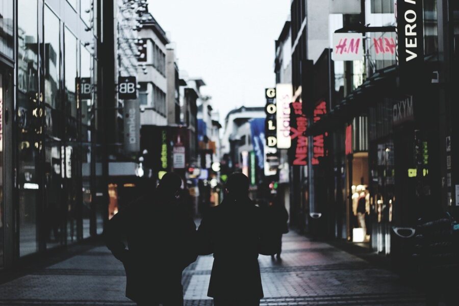 Two people walk down a dimly lit urban street lined with shops and illuminated signs at dusk.