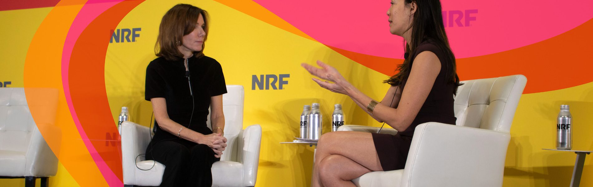 Two women seated on stage engage in conversation. One gestures while speaking, and the other listens. Bright yellow and pink background with "NRF" logo visible.