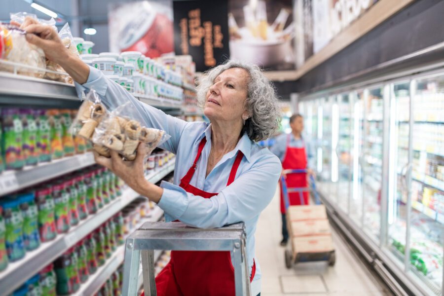 An older person in a red apron, standing on a ladder, is stocking shelves with packaged goods in a grocery store aisle.
