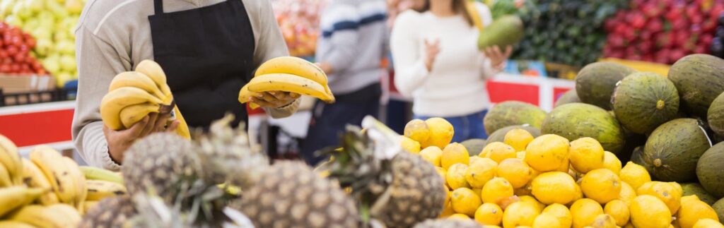 A person in an apron holds bananas at a fruit market. Pineapples, lemons, and avocados are displayed. Two people in the background examine produce.