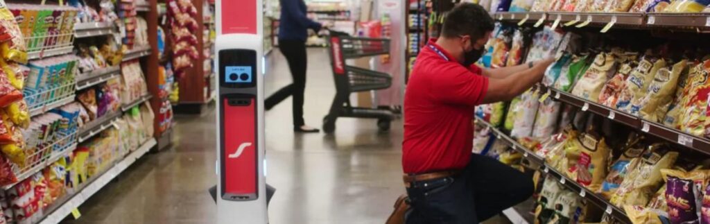 A person in a red shirt and mask kneels to stock shelves in a grocery aisle filled with chips. A robot stands nearby. Another person shops in the background with a cart.