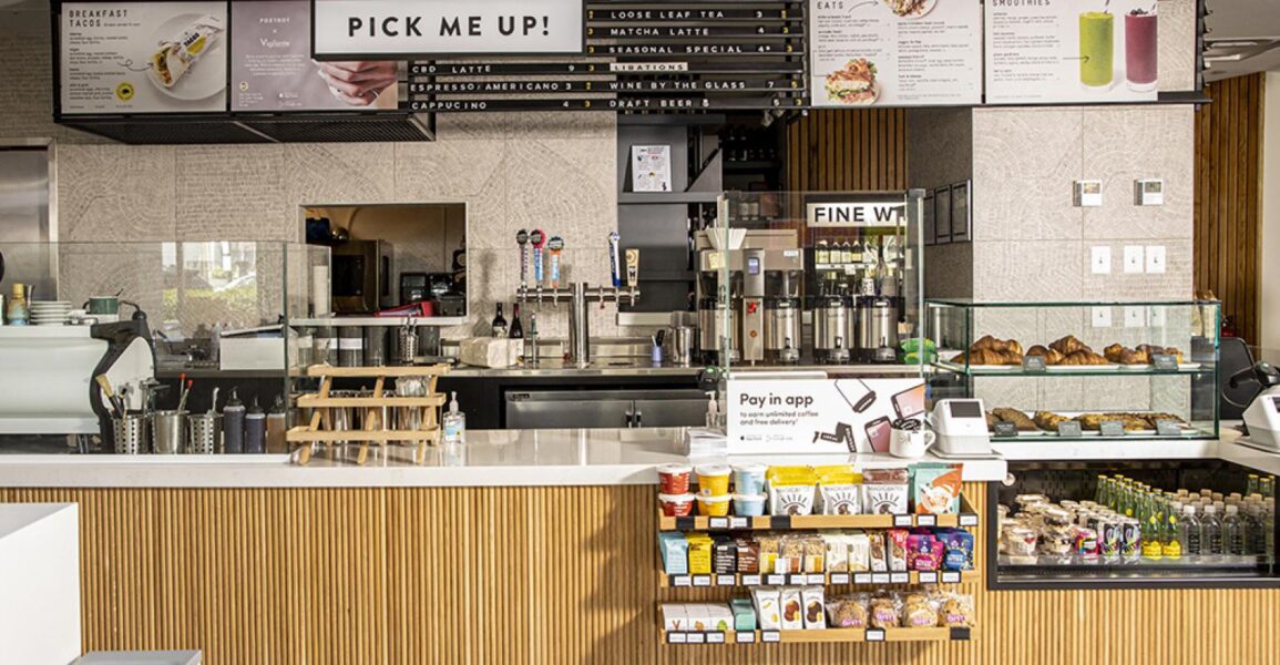 Counter service cafe with a wooden panel counter, menu above, various pastries in a glass case, snacks on display, and a payment station. Coffee machines and drink options are visible in the background.