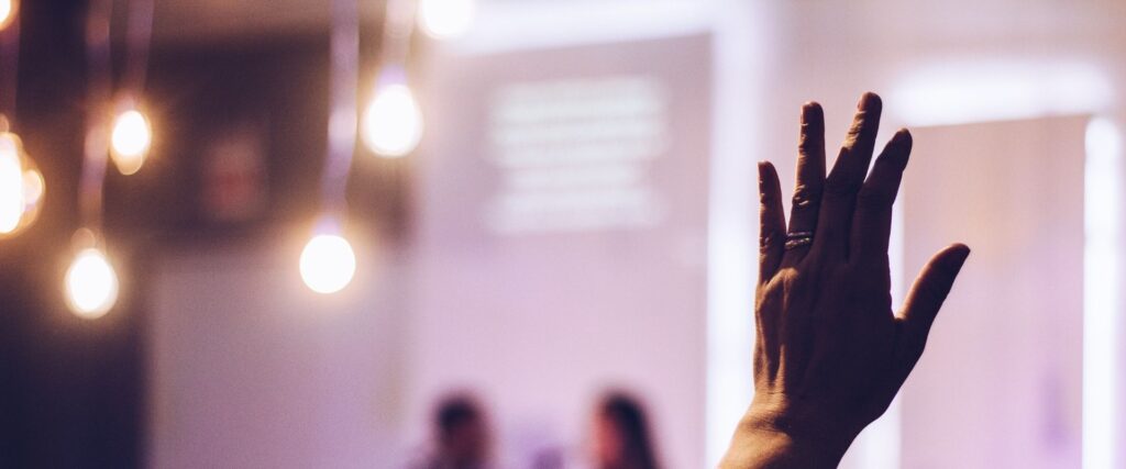 A raised hand in a room with blurred figures in the background and hanging light bulbs.