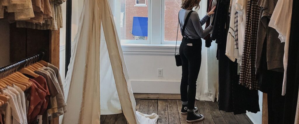 Person browsing clothes in a boutique store, standing near a fitting room with natural light coming in through a window.