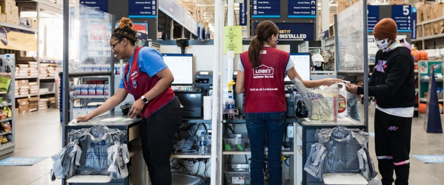 Two checkout counters in a store with employees in red vests assisting customers. Plastic barriers are between the stations, and bags of purchases are on the counters.