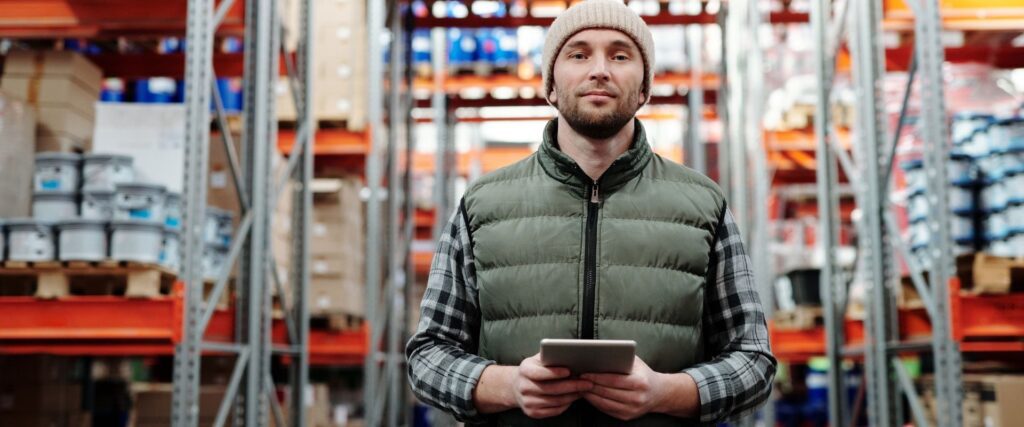 Man wearing a beanie and green vest holds a tablet in a warehouse with shelves of boxes and supplies.