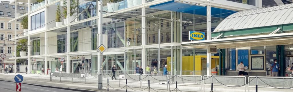 Glass-fronted IKEA store with multiple levels and trees on balconies, set on a bustling city street with pedestrians and a bike lane.