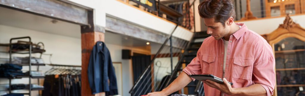 A person in a pink shirt uses a tablet while standing in a clothing store with racks and shelves in the background.