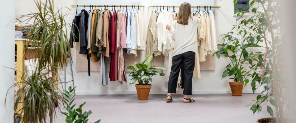 A person with long hair, wearing a white shirt and dark pants, browses a rack of assorted-colored clothes in a store, surrounded by potted plants.