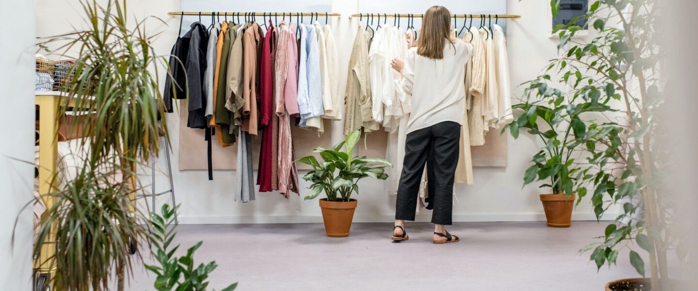 A person with long hair, wearing a white shirt and dark pants, browses a rack of assorted-colored clothes in a store, surrounded by potted plants.
