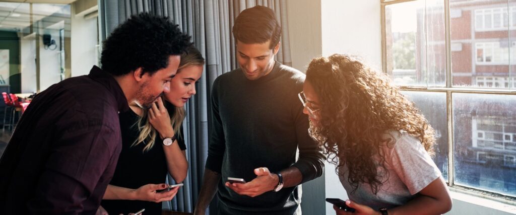 Four people stand in a well-lit room, looking at a smartphone.