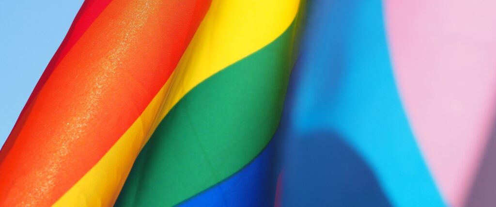 Close-up of rainbow flag and transgender pride flag waving against a clear blue sky.