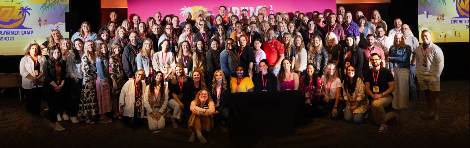 A large group of people poses together indoors in front of a colorful "Summer Camp 2023" backdrop.