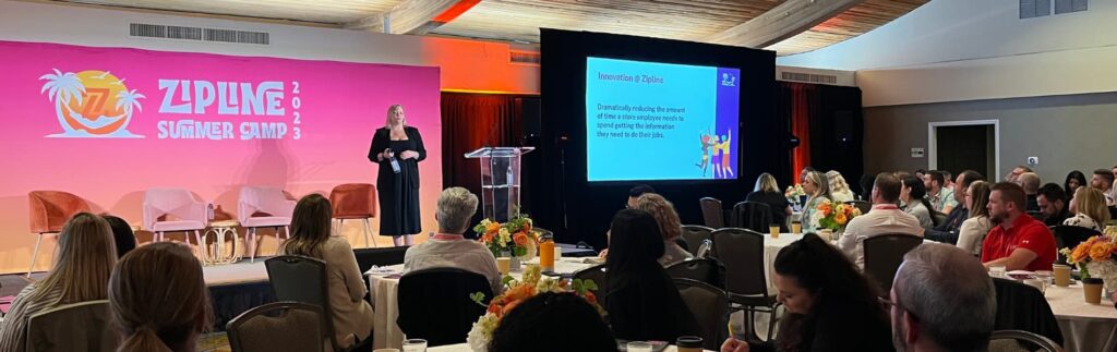 A person speaks on stage at a conference with a screen displaying a presentation titled "Innovation in Crisis." The backdrop reads "Zipline Summer Camp 2023." Audience members are seated.