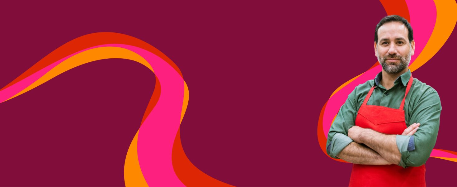 Man with a beard wearing a red apron stands with arms crossed against a colorful abstract background.