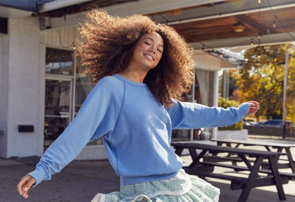 Person with curly hair wearing a blue sweatshirt and floral skirt spins outdoors near a wooden picnic table, with a building and autumn trees in the background.