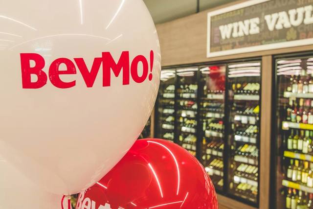 White and red balloons with "BevMo!" text in front of refrigerated wine displays labeled "Wine Vault" in a store.