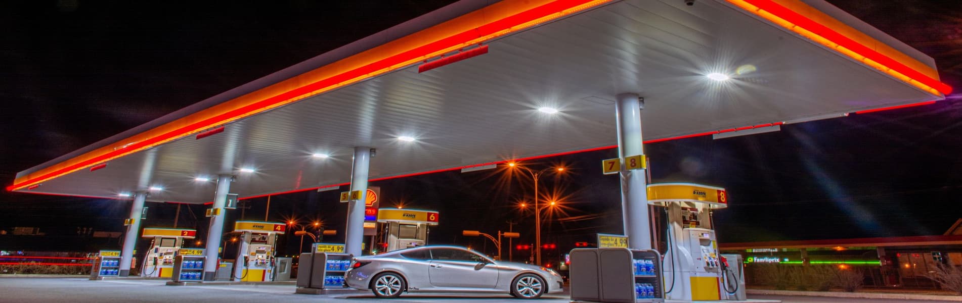 A car is parked at a brightly lit gas station at night, with overhead lights illuminating four fuel pumps.