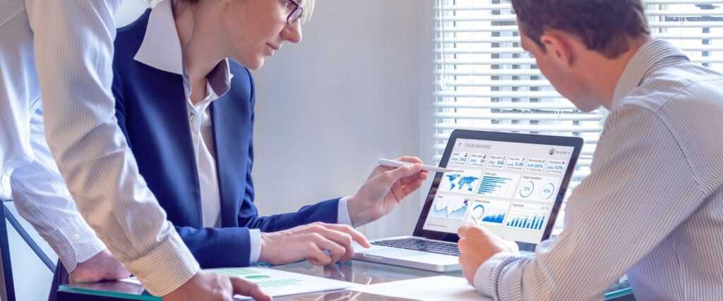 Three people in a meeting room review analytics on a laptop displaying various charts and graphs. One person points at the screen with a pen, discussing the data.