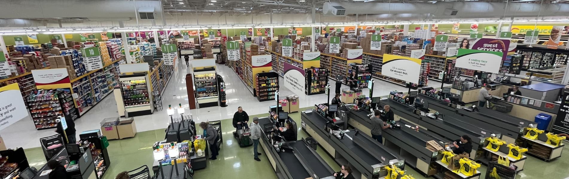 Aerial view of a spacious supermarket interior with aisles and checkout counters. Shoppers and employees are visible. Bright lighting and signage indicate various sections.