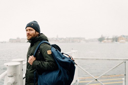 A man with a beard stands on a dock carrying a large blue backpack, wearing a gray knit hat and green jacket. The background shows an overcast sky and a body of water.
