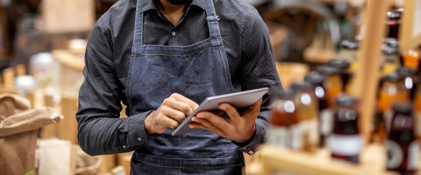 Person wearing a dark shirt and blue apron, using a tablet in what appears to be a store with bottles on a shelf in the background.