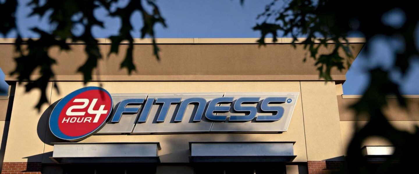 Sign on a building for "24 Hour Fitness," featuring a large logo and stylized text.