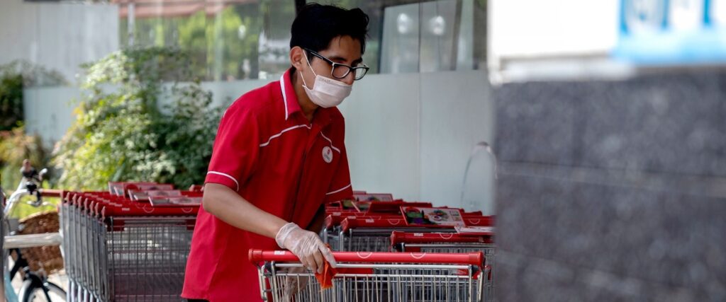 A person wearing a red shirt, mask, and gloves pushes a row of shopping carts outdoors.