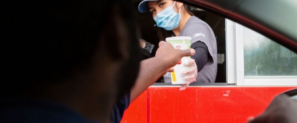 Drive-thru employee hands a cup to a customer in a car. The employee wears a mask and a cap.