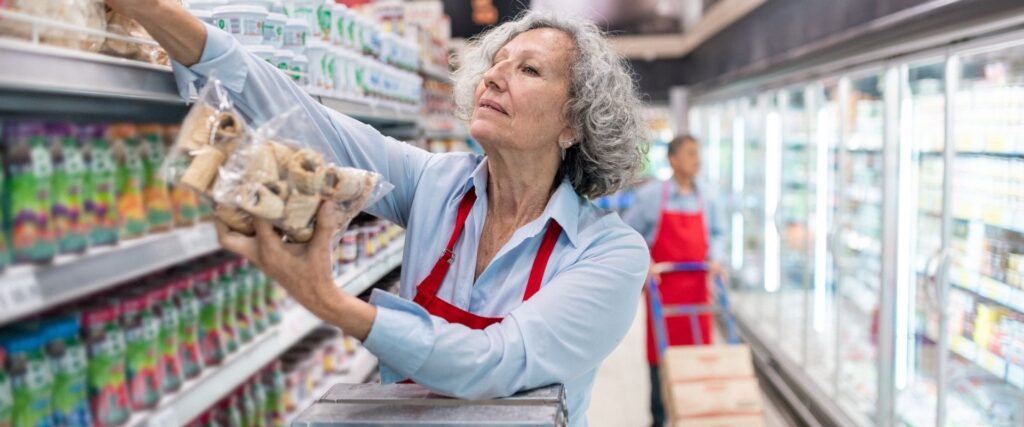 An older woman wearing a red apron and blue shirt stocks shelves with bagged products in a grocery store aisle. Another person in a red apron stands in the background near a cart.