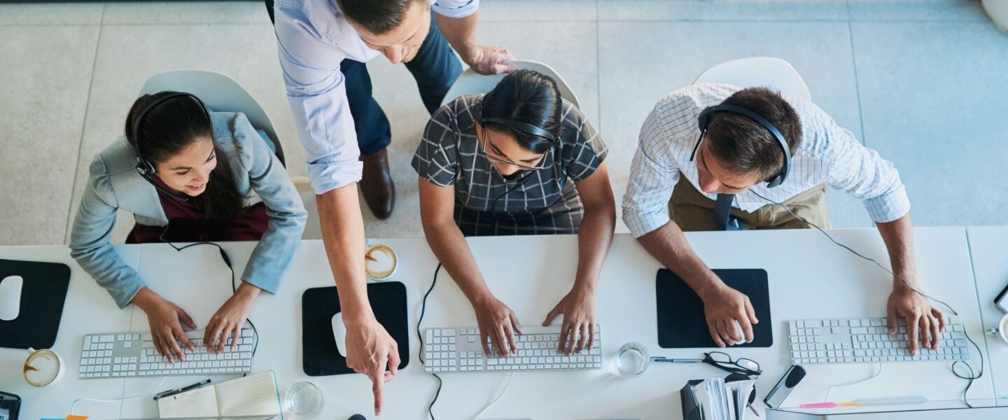 Four people working at computers, one standing and pointing at a screen, while the others sit with headsets, viewed from above.