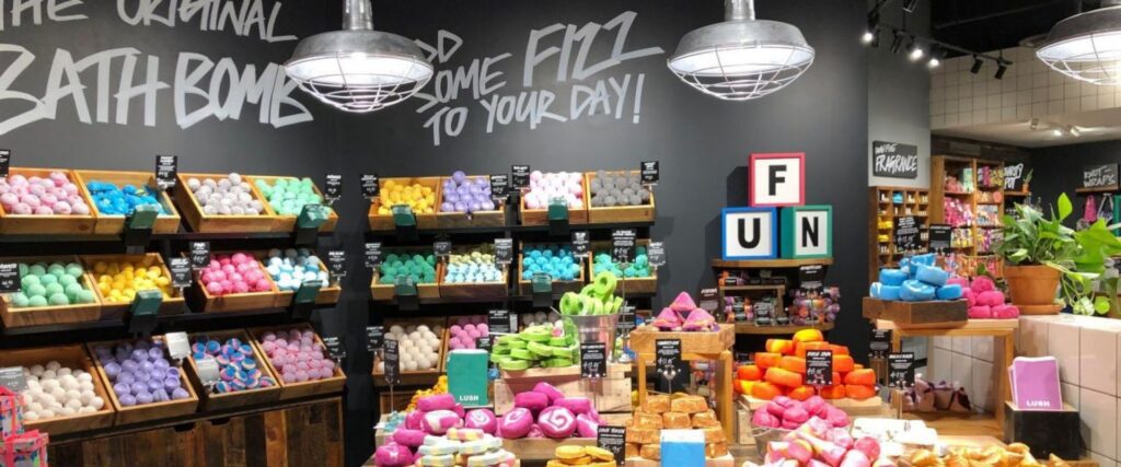 A brightly lit store displays a variety of colorful bath bombs on wooden shelves, with playful signage on the walls and ceiling lights above.