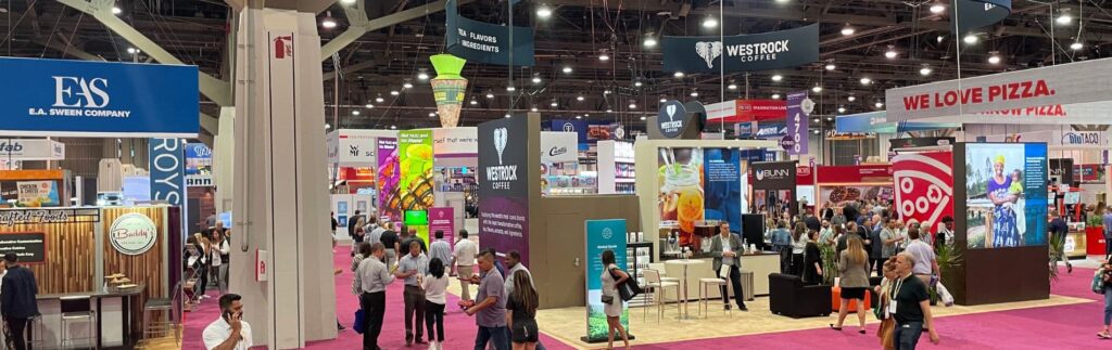 A crowded trade show floor with various booths, banners, and attendees. Bright lighting and colorful displays are present.