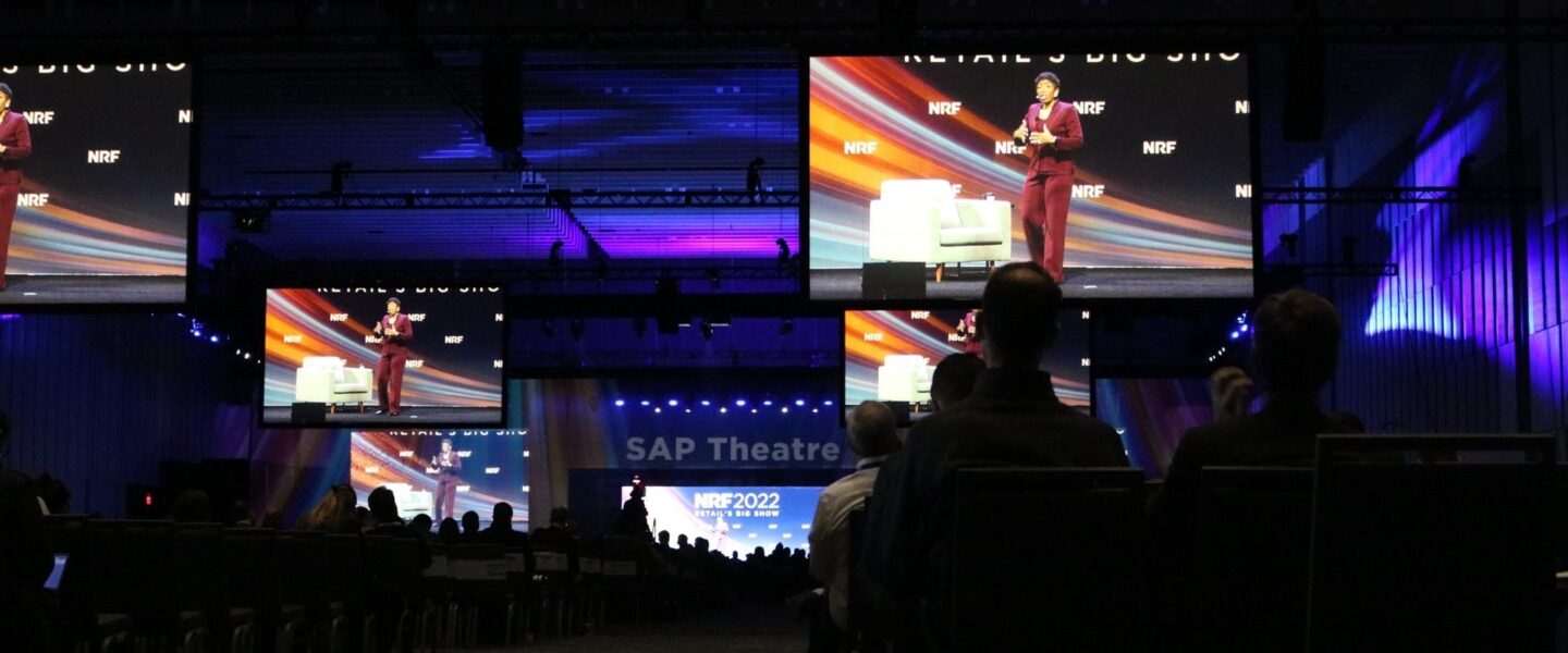 A speaker presents on stage at the NRF 2022 event in a dark auditorium, with multiple large screens displaying the presentation.