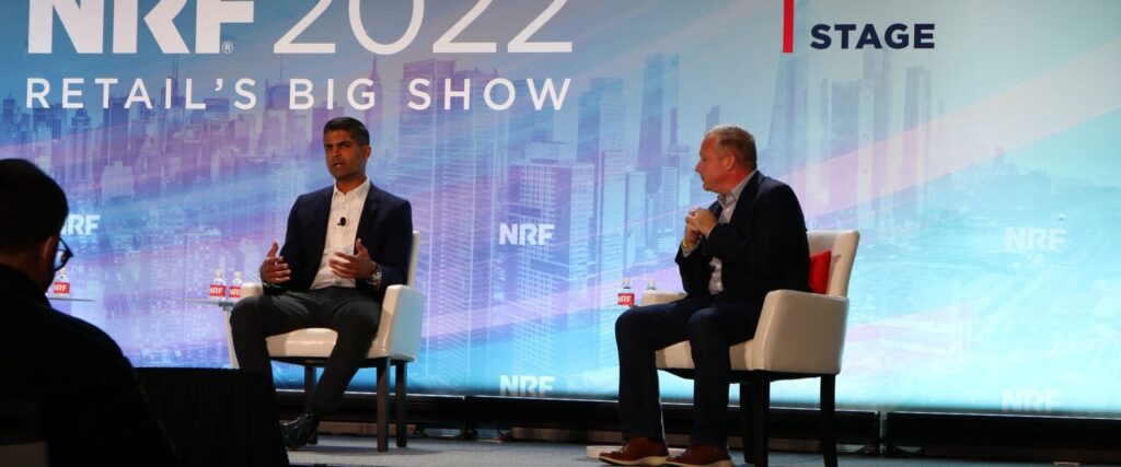 Two men sit in chairs on a stage during the NRF 2022 Retail's Big Show, engaged in a discussion. The backdrop features the event's branding.
