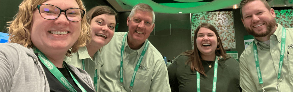Five people smiling, wearing lanyards and similar shirts, standing indoors at an event.