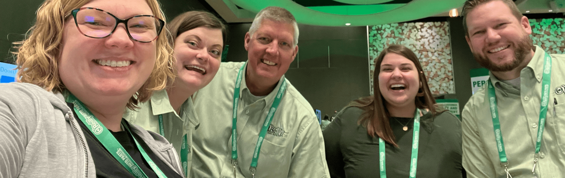 Five people smiling, wearing lanyards and similar shirts, standing indoors at an event.