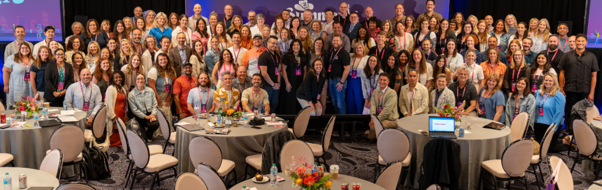 A large group of people standing together in a conference room, posing for a group photo. Round tables with flowers and conference materials are in the foreground.