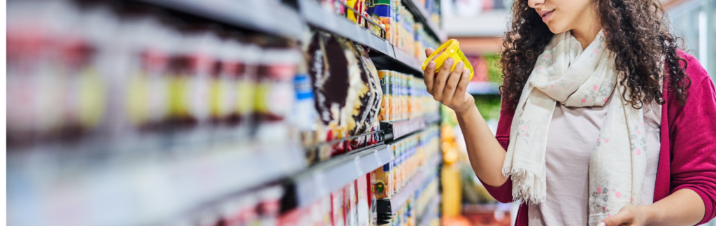 Person shopping in a grocery store, holding and examining a yellow packaged item from a shelf filled with various products.