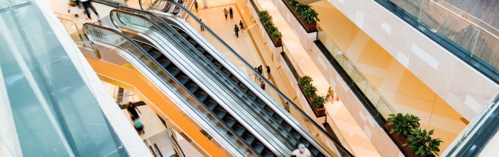 Overhead view of escalators in a shopping mall with people and plants nearby.