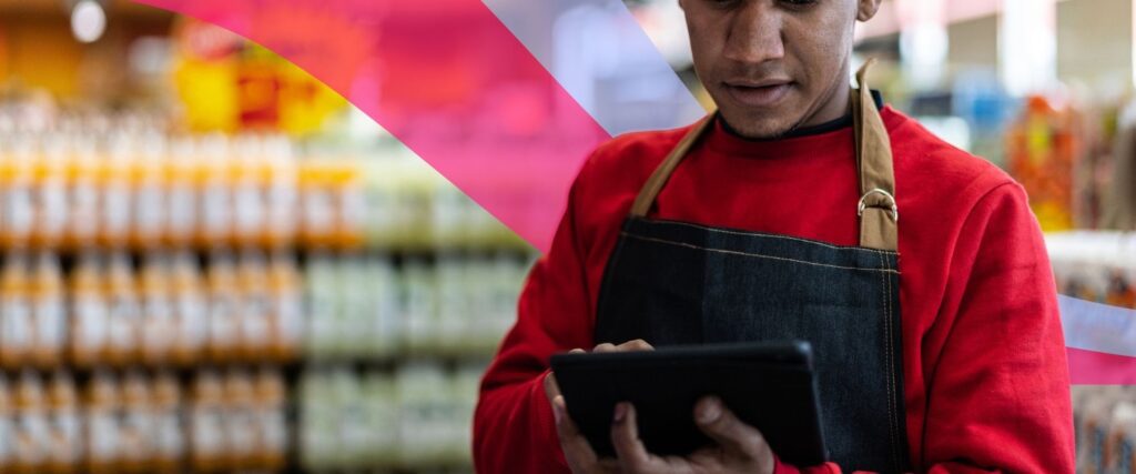Store employee in a red shirt and apron uses a tablet, with shelves of products visible in the background.