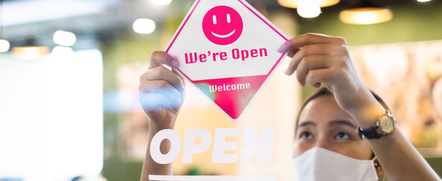 A person wearing a mask is placing an "Open" sign with a smiling face on a glass door.