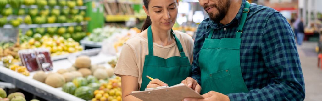 Two workers in green aprons review a clipboard in a grocery store's produce section, surrounded by various fruits and vegetables.