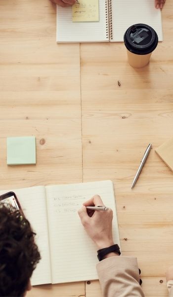 Two people working at a wooden table with notebooks, a pen, sticky notes, and a coffee cup.